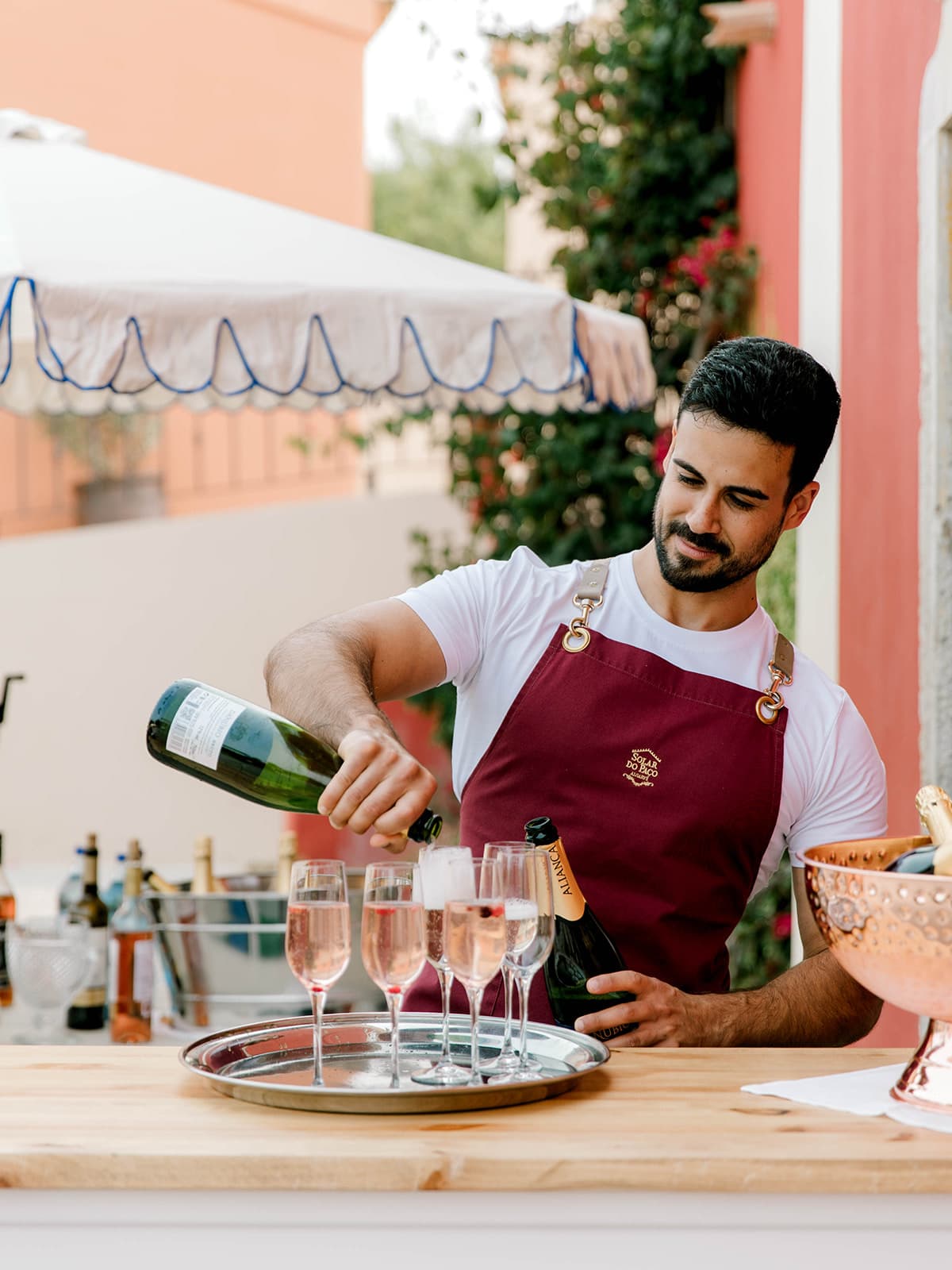 A barman in a burgundy apron pouring pink sparkling wine into glasses at a drinks station, Solar do Paço Algarve