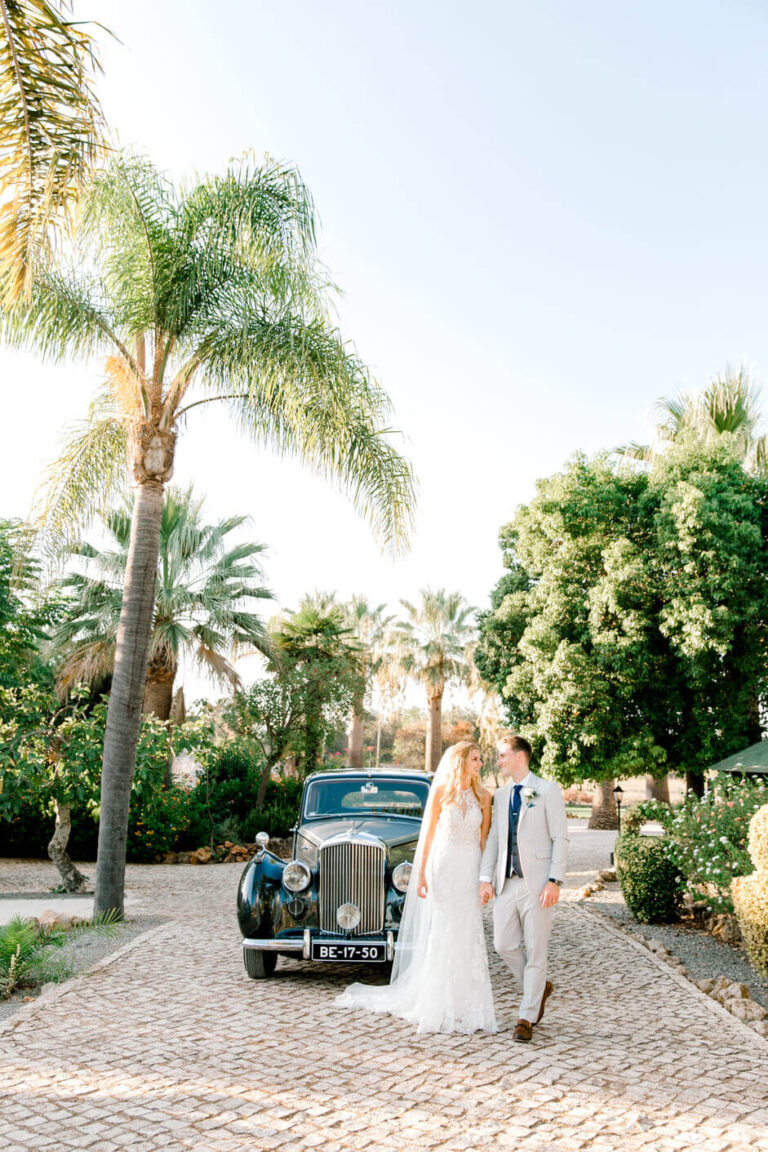 Bride and groom walking beside a vintage car among palms, Sonho a Dois Algarve Wedding Planners