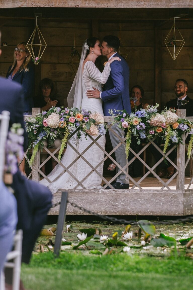 The couple kiss beneath a timber and foliage arch, Sorcha Herbert Flowers