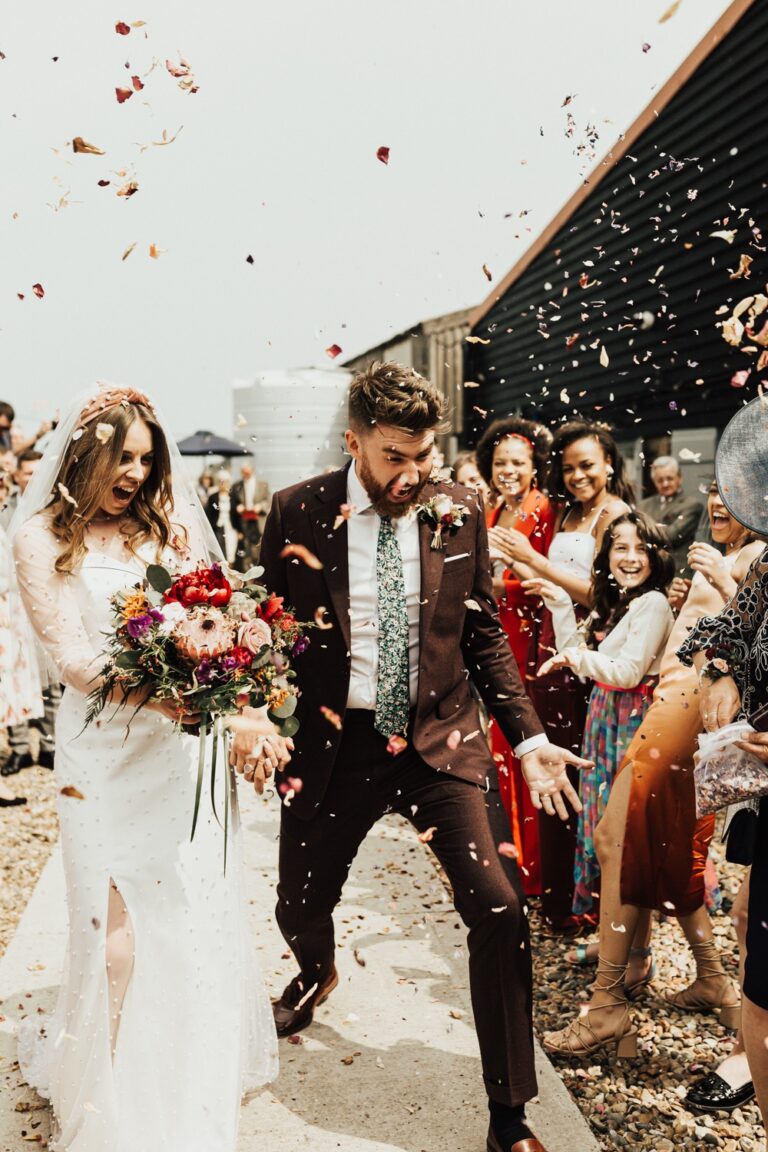 Running through confetti, the wedding party behind them, Sorcha Herbert Flowers
