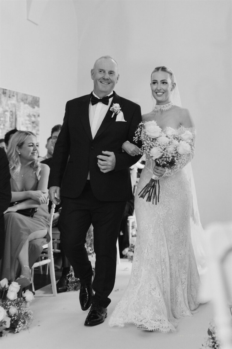 The bride's father walks her down the aisle, her bouquet of white roses and gypsophila in hand, Sparx Wedding & Event Planner