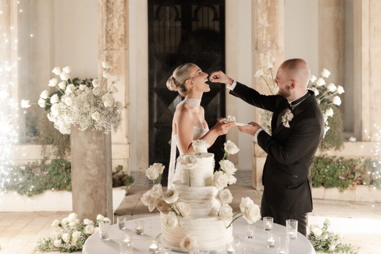 The couple feeding each other cake as sparklers catch, beside a tiered white cake dressed in roses, Sparx Wedding & Event Planner