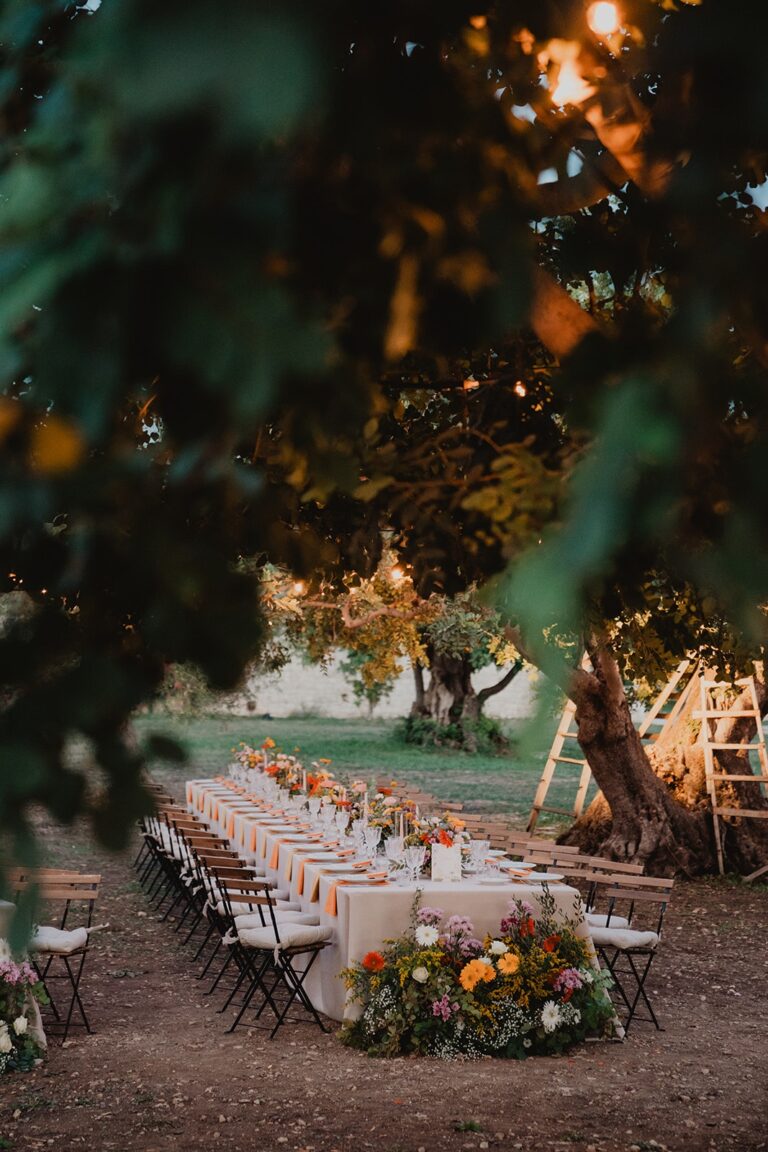 A long table laid for dinner beneath the trees at dusk, orange and gold florals running its length, Sparx Wedding & Event Planner
