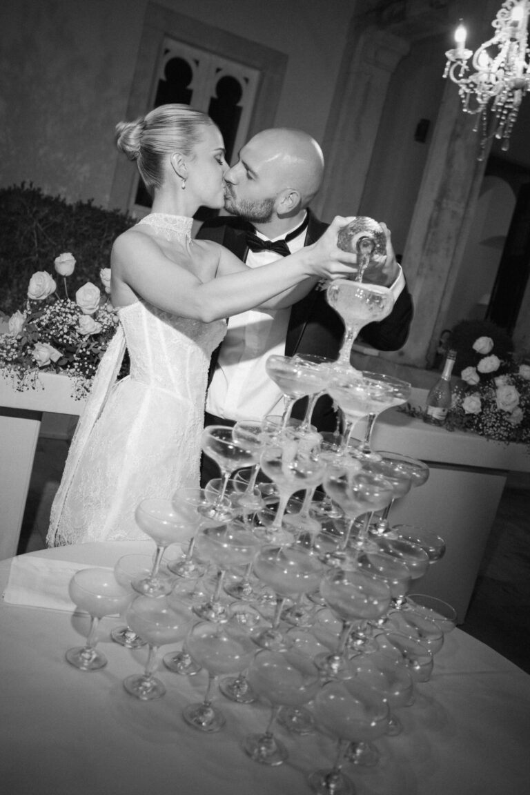 The newlyweds kiss beside a champagne tower, coupe glasses stacked in tiers on the table, Sparx Wedding & Event Planner