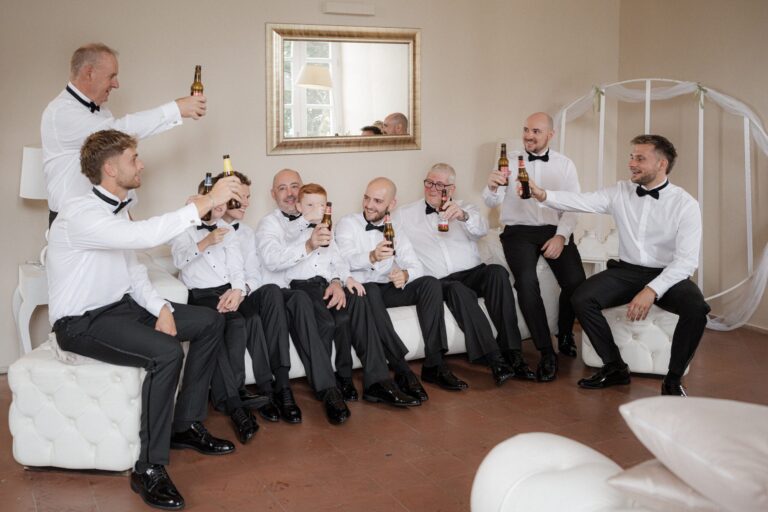 Groomsmen raise bottles of beer together, mid-toast on a white sofa before the wedding, Sparx Wedding & Event Planner