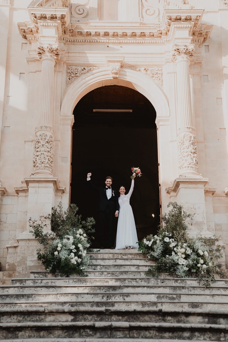 The newlyweds cheering on the cathedral steps in Noto, the bride's bouquet raised overhead, Sparx Wedding & Event Planner