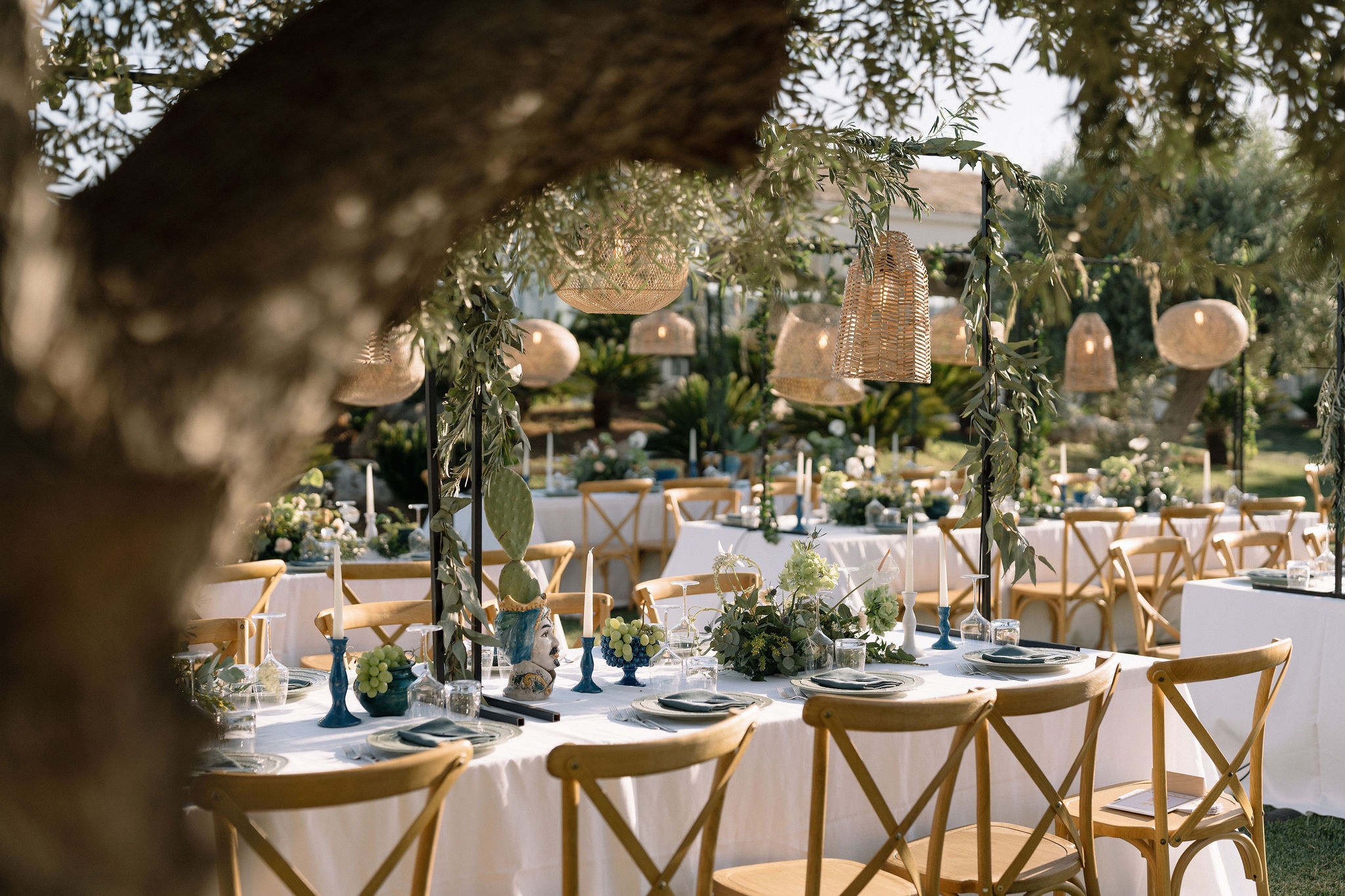 A Sicilian ceramic head vase and market baskets of grapes and citrus, set beneath olive trees strung with woven lanterns, Sparx Wedding & Event Planner