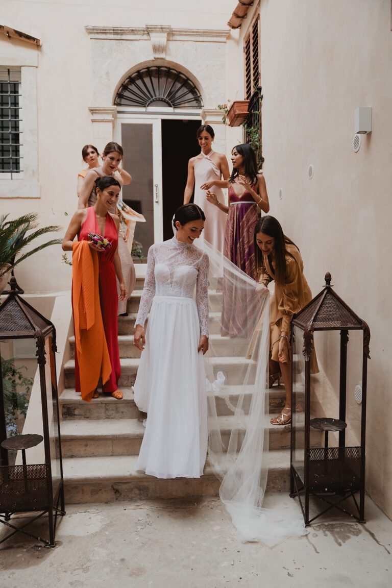 Bridesmaids help the bride down a staircase, her veil trailing the stone steps behind her, Sparx Wedding & Event Planner