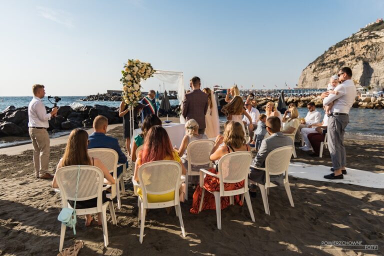 A beach ceremony: a floral arch on the sand, cane chairs, the headland behind, Sposa Mediterranea By A&C