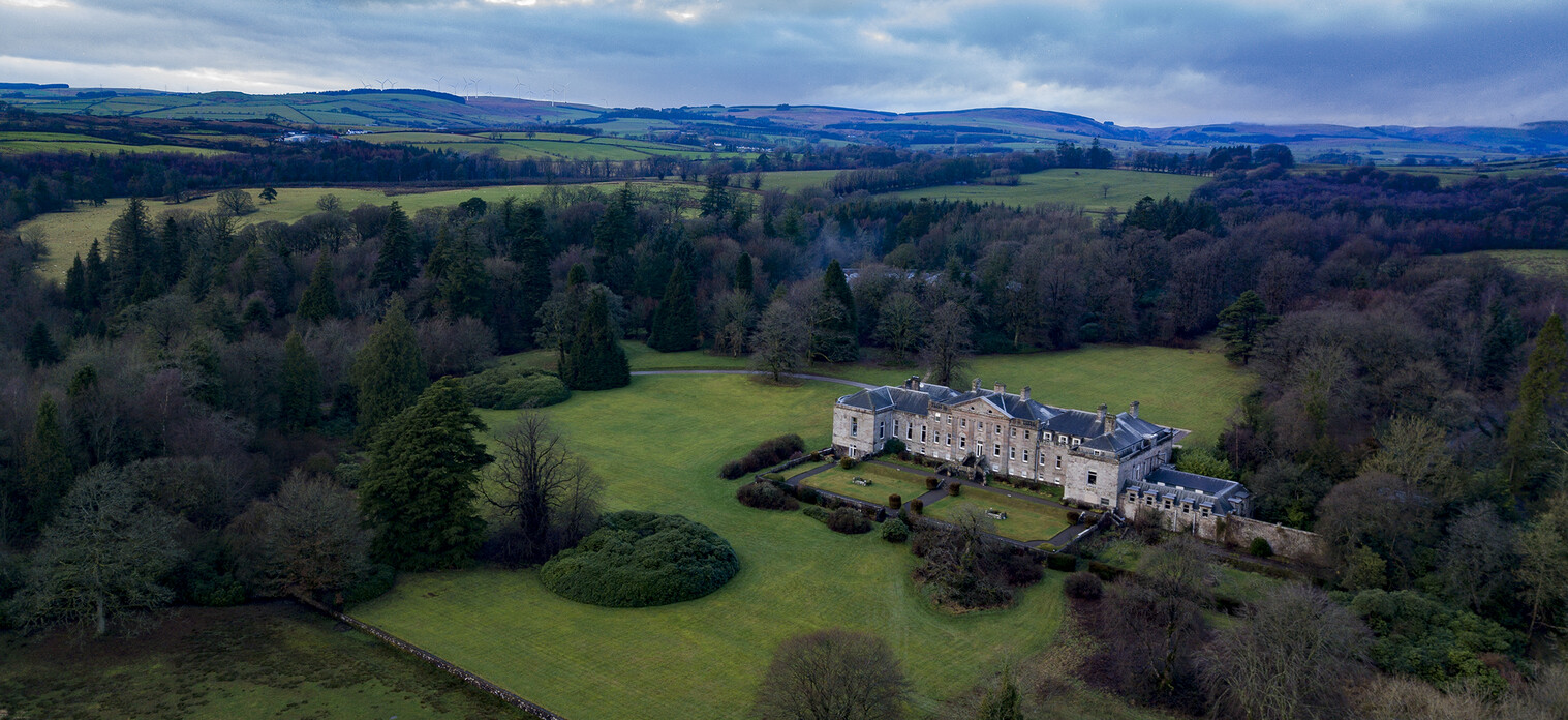 A wider aerial view showing the house in a clearing of lawns and mixed woodland, with the Dumfriesshire hills beyond, Springkell