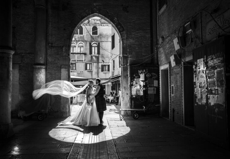 A veil lifting in the wind as a couple pause under a Venetian archway, in black and white, Stefano Lunardi