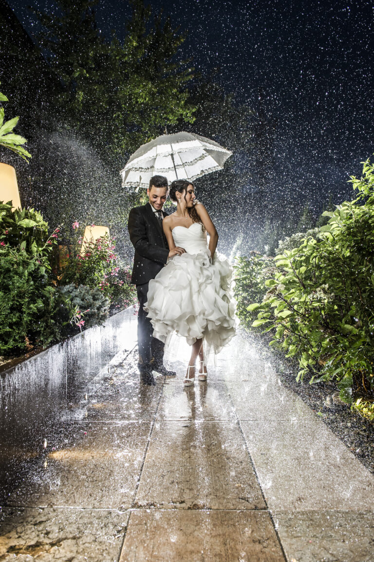 Newlyweds under a white umbrella on a garden path in heavy evening rain, Stefano Lunardi