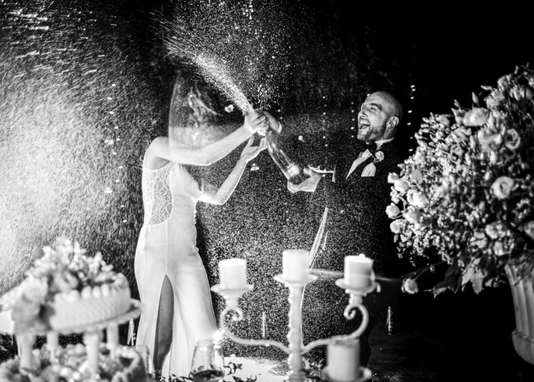 Champagne spraying as the couple open a bottle beside the cake at night, in black and white, Stefano Lunardi