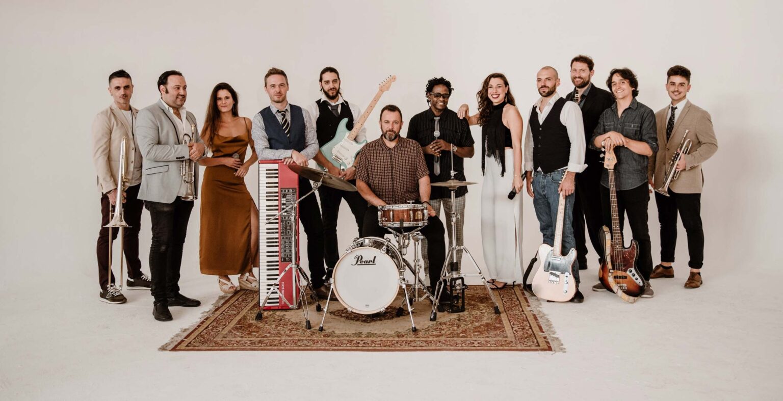 The full fourteen-piece band lined up on a patterned rug in a white studio