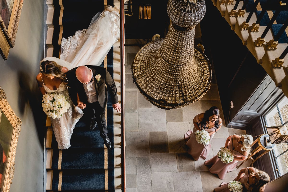 A bride on the staircase seen from above, with bridesmaids waiting in the hall below
