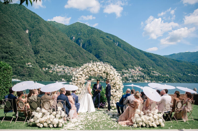 A ceremony under the white rose arch on Lake Como, guests holding white parasols, Suellen Fernandes Weddings & Celebrations