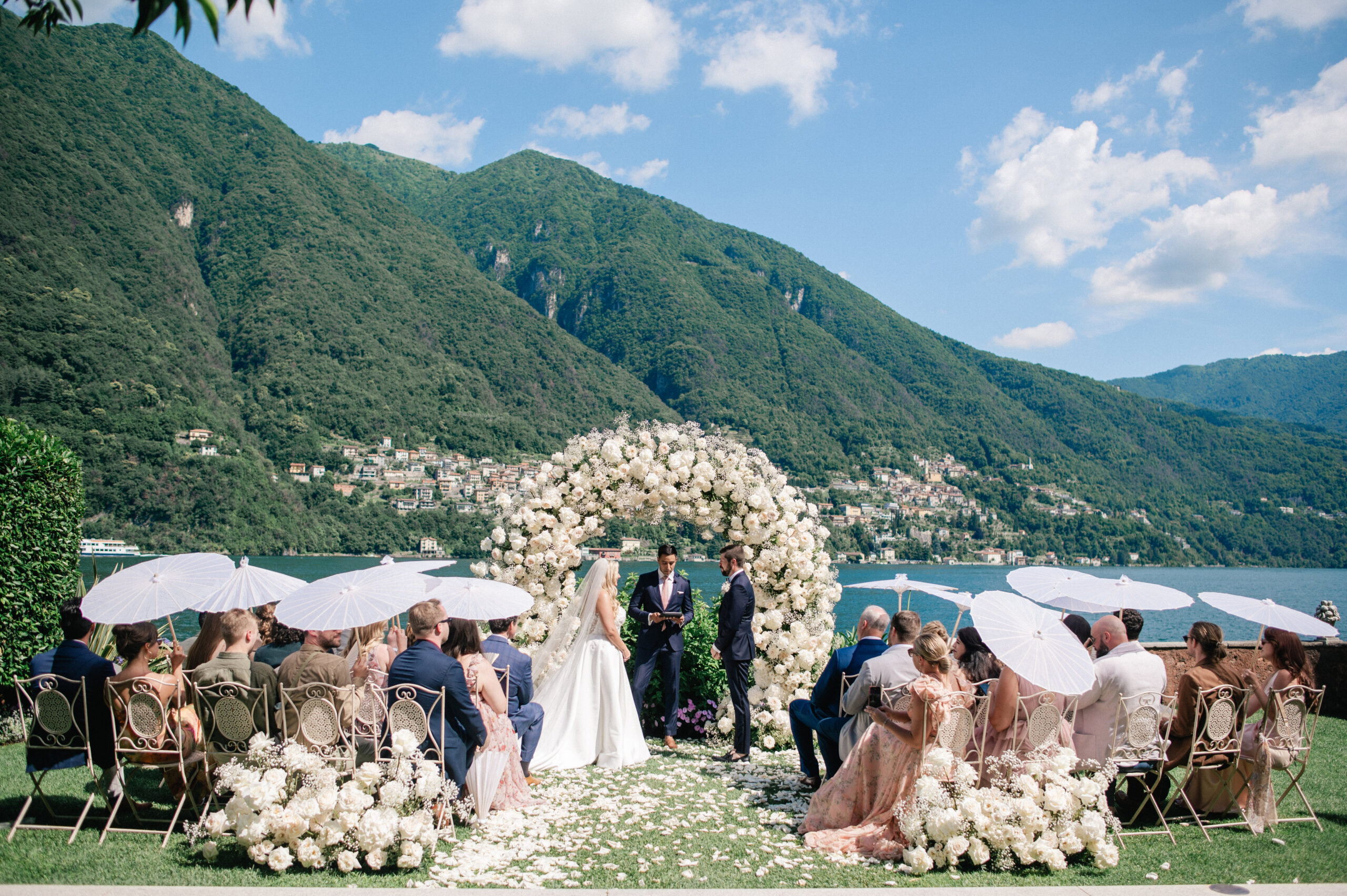 A ceremony under the white rose arch on Lake Como, guests holding white parasols, Suellen Fernandes Weddings & Celebrations