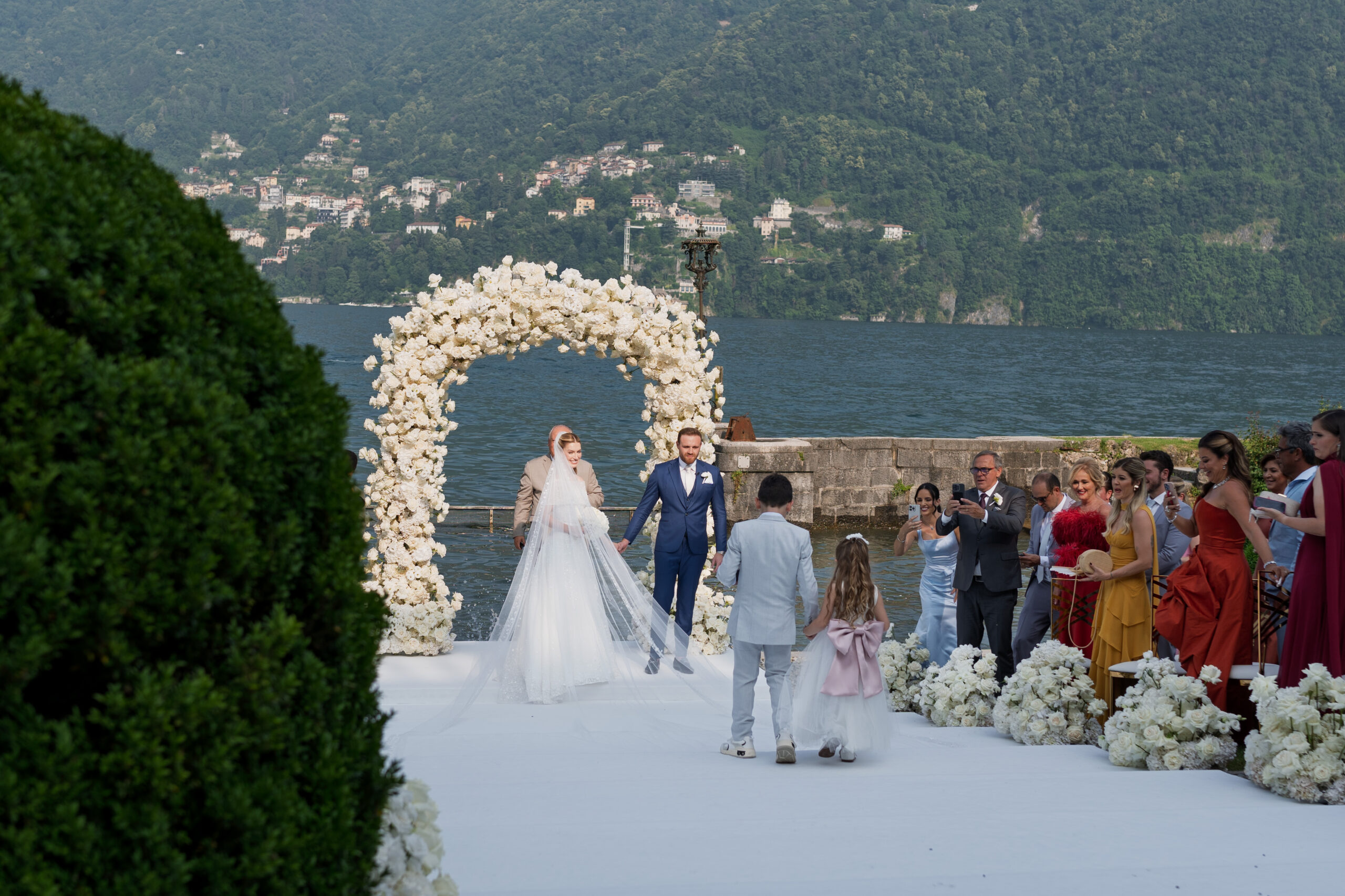 A ceremony on a white aisle at the water's edge of a Lake Como villa, flower girl and guests either side, Suellen Fernandes Weddings & Celebrations