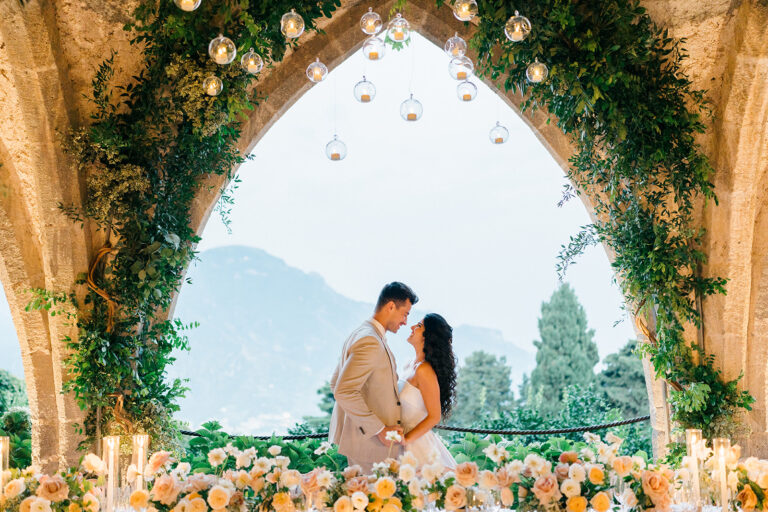 The couple under a gothic arch hung with greenery and glass lanterns, coastline beyond, Ravello, Suellen Fernandes Weddings & Celebrations