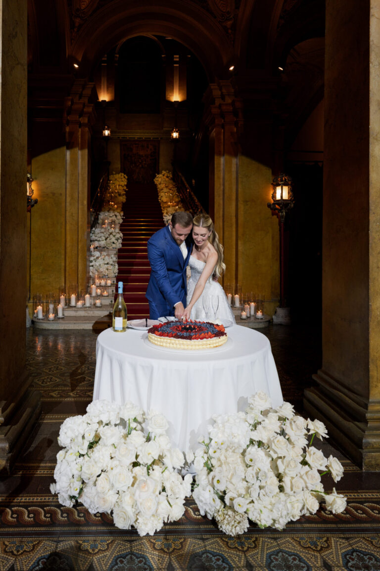 Cutting the cake at the foot of a palazzo staircase, the table banked with white flowers, Suellen Fernandes Weddings & Celebrations