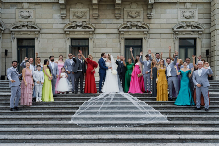 The wedding party on the steps of a Lake Como villa, arms raised, the veil spread down the stairs, Suellen Fernandes Weddings & Celebrations