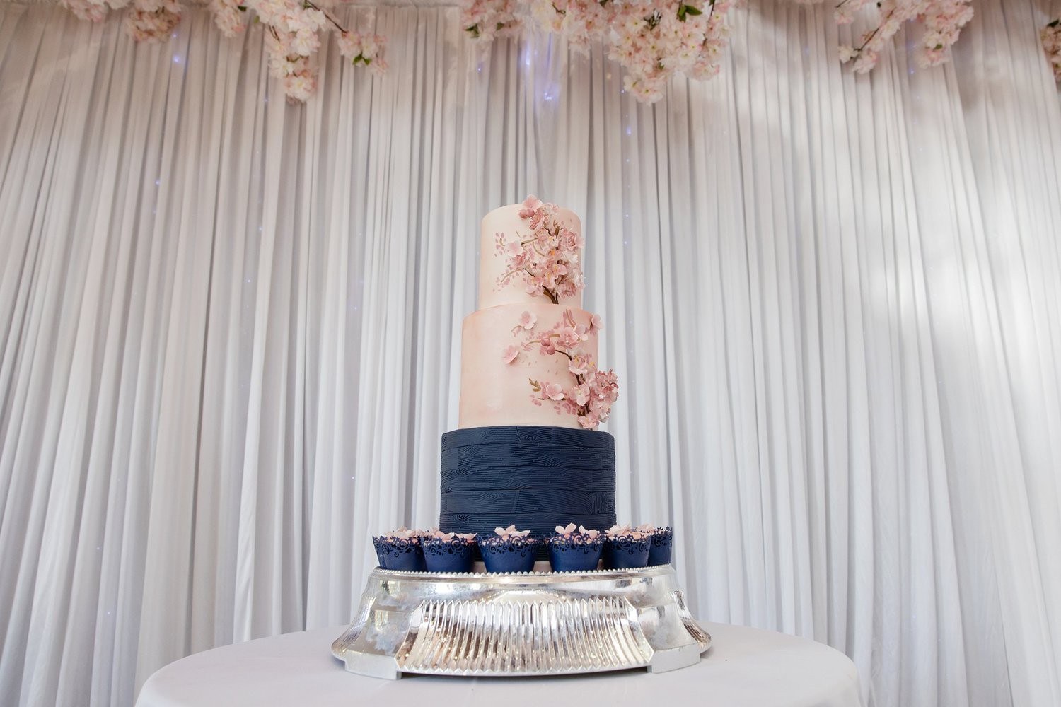 A navy and white tiered cake with pink sugar flowers and matching cupcakes, Sugar and Spiced Cakes