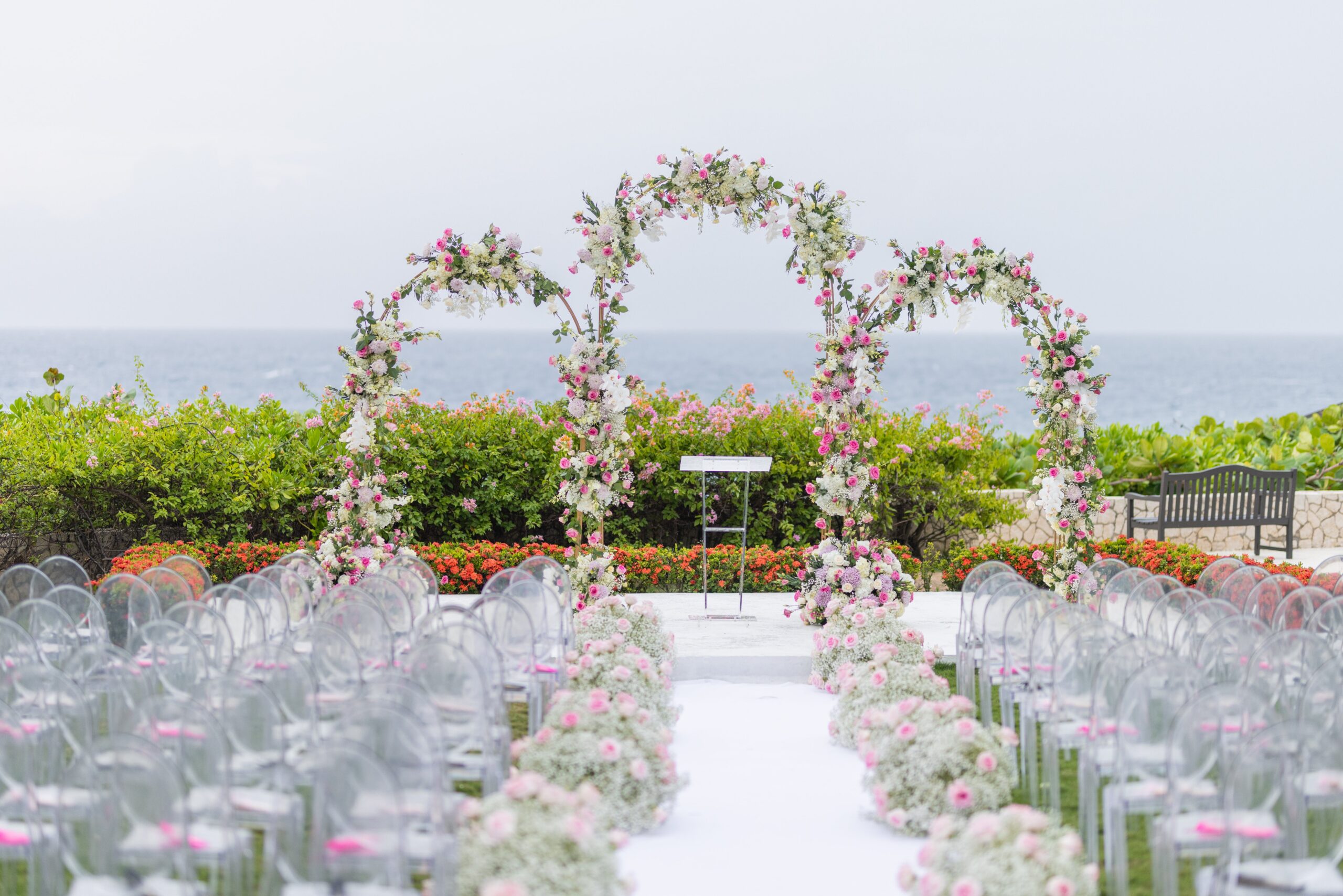 Three round floral arches over a white aisle lined with flowers and clear chairs, above the sea at The Cliff Hotel, Tai Flora Luxe