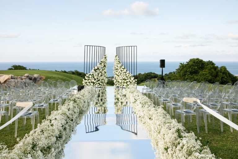 A mirrored aisle between banks of white flowers and rows of chairs, with a tall gate and the sea behind, Tai Flora Luxe