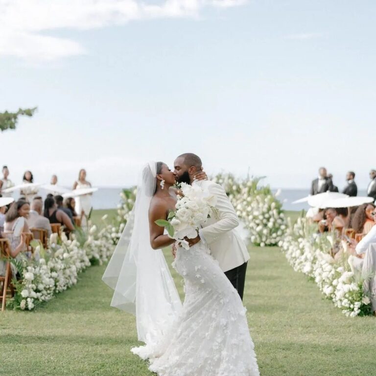 A couple kissing in the aisle, lined with white floral borders, with the sea behind, Tai Flora Luxe