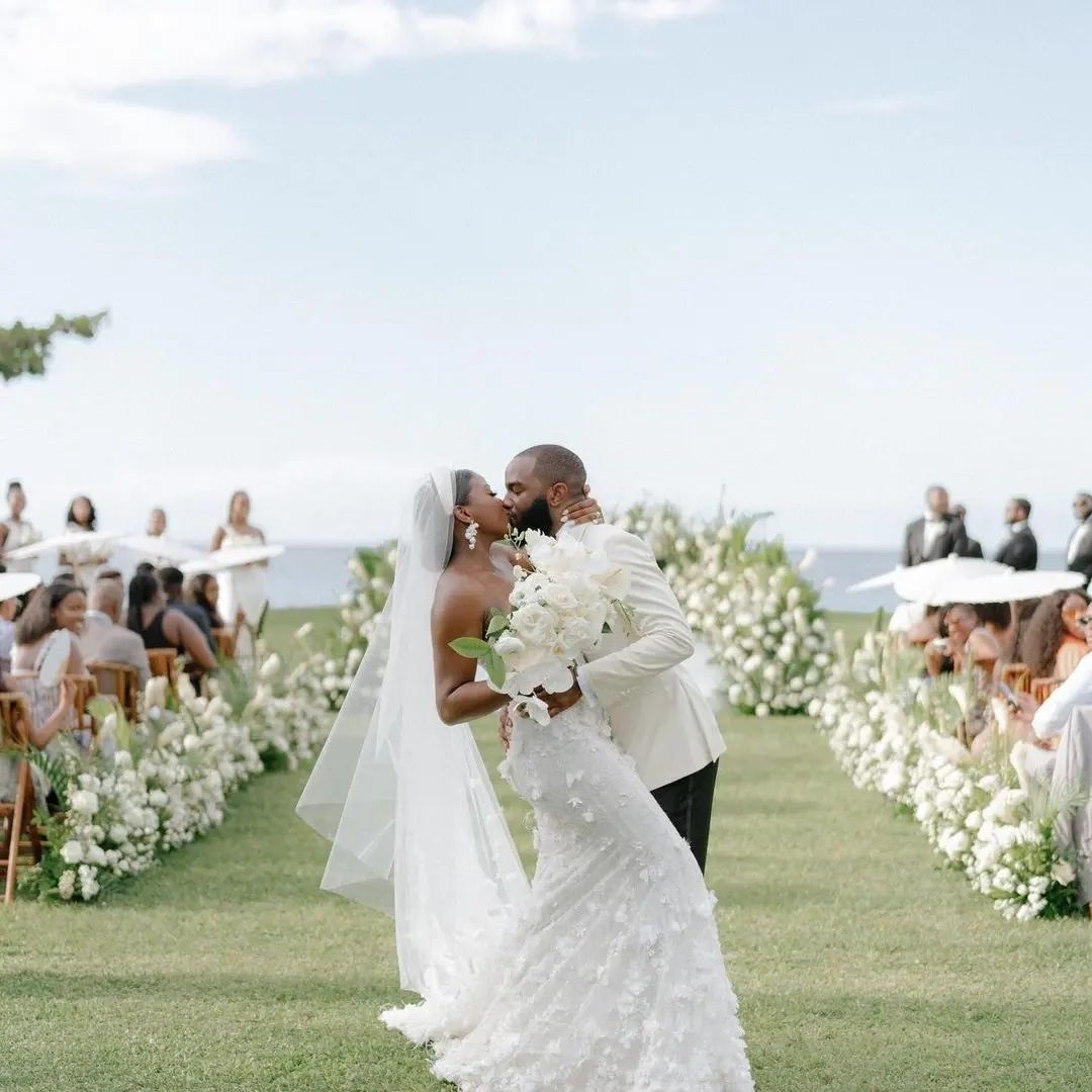 A couple kissing in the aisle, lined with white floral borders, with the sea behind, Tai Flora Luxe