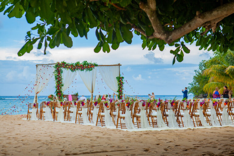 A beach ceremony with a draped, flower-garlanded canopy and rows of wooden chairs facing the shore, Tai Flora Luxe