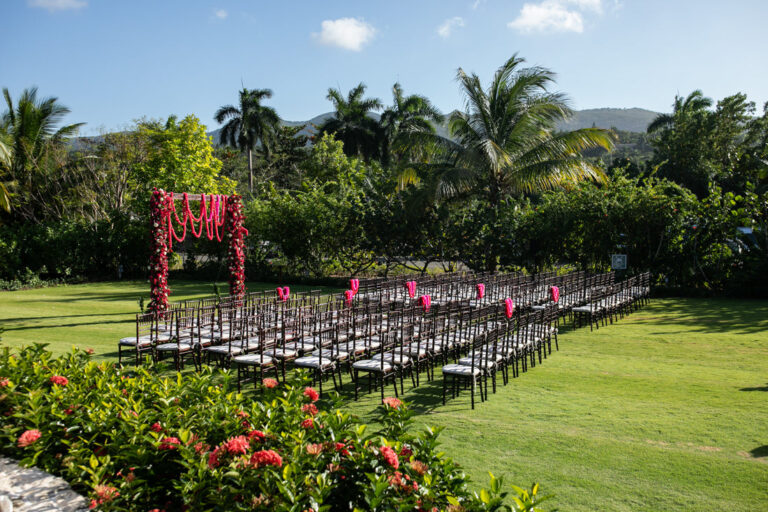 Rows of dark chairs on a manicured lawn leading to a pink floral arch at Half Moon, Montego Bay, Tai Flora Luxe