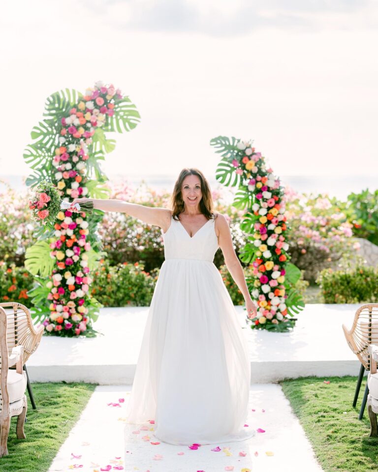 A bride holding a bouquet between two tall columns of pink, orange, yellow and white roses with monstera leaves, Tai Flora Luxe