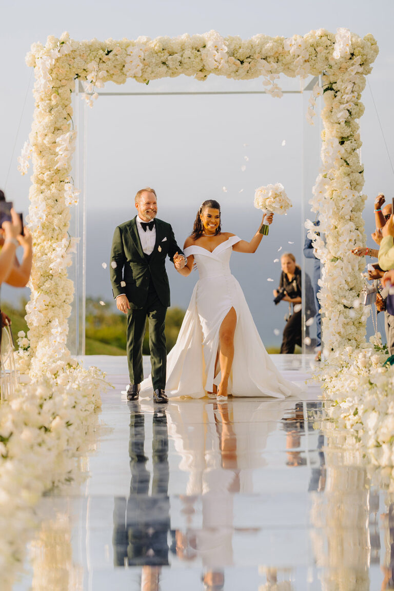 A bride and groom walking through a large mirrored frame edged with cream flowers, Tai Flora Luxe
