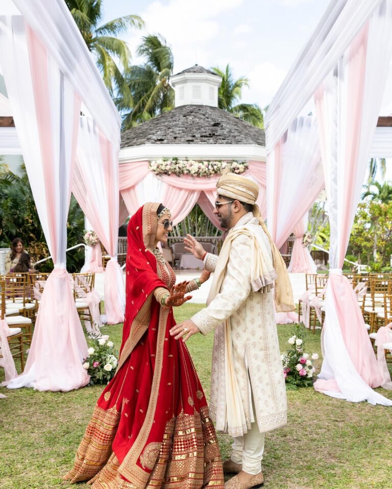 A couple in red and ivory South Asian wedding dress beside a canopy of pink drapes, Tai Flora Luxe