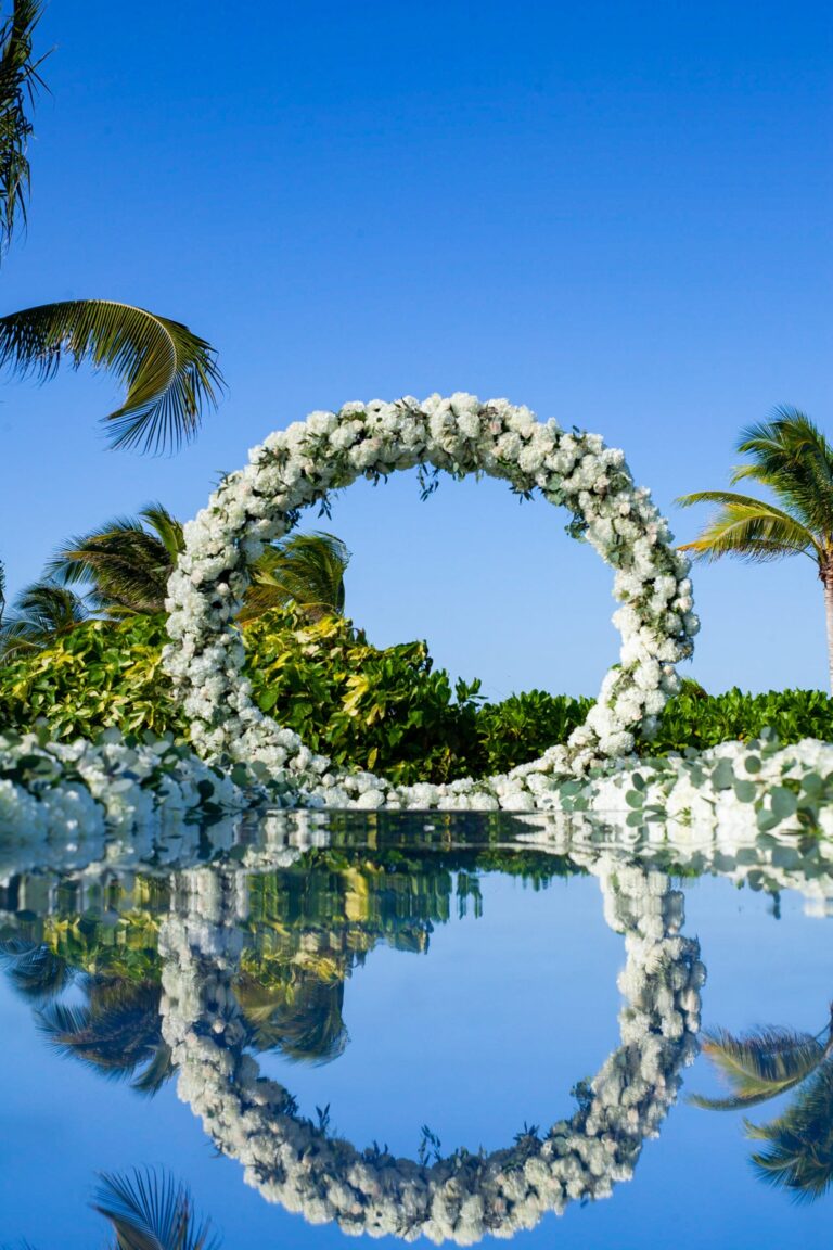 A circular floral ring beside a pool, reflected in the water against a blue sky, Tai Flora Luxe