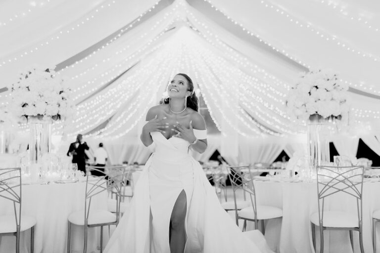 A bride laughing beside tables with tall white centrepieces under a white tent, in black and white, Tai Flora Luxe