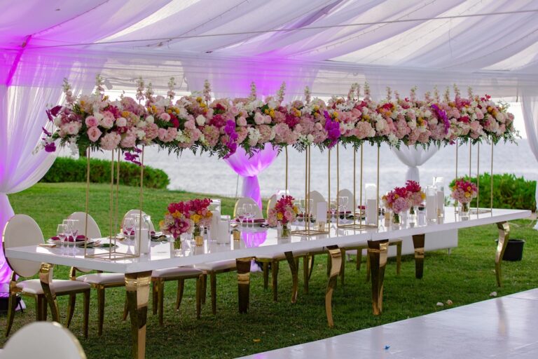A long reception table under a lit tent, with a low floral garland in pink, white and purple, Tai Flora Luxe