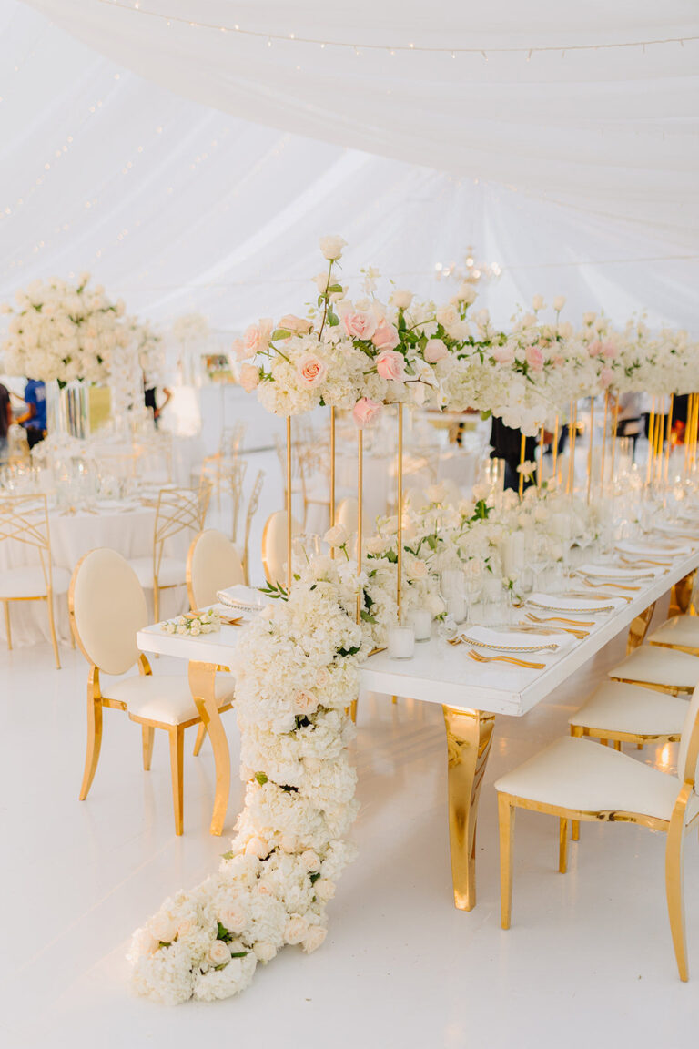 A long white table with a runner of cream and blush flowers and gold chairs under a draped tent, Tai Flora Luxe