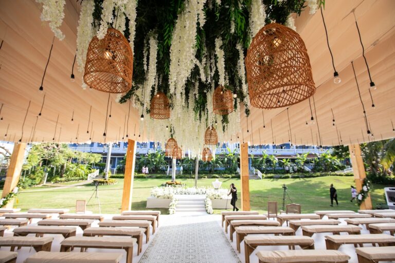Hanging white blooms and woven pendant lights over ceremony seating in an open-sided tent on a lawn, Tai Flora Luxe