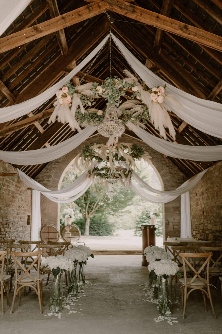The arched stone barn set for a ceremony, with draped fabric, a chandelier and white flowers along the aisle, Tan House Newland