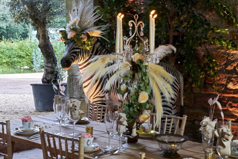 A table set with crystal candelabra, pampas plumes and a zebra model beside an olive tree, Tan House Newland