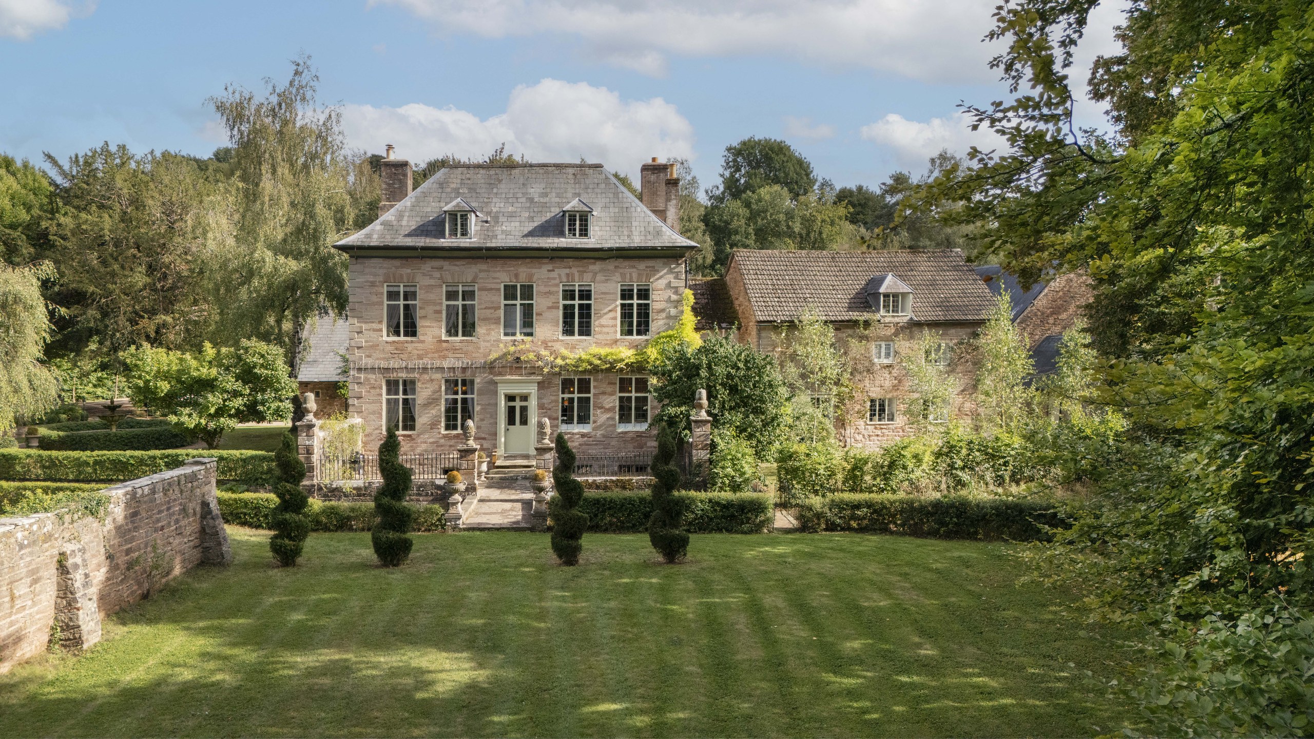 The stone house seen across its lawn, with clipped yews, a low garden wall and the old outbuildings to the right, Tan House Newland