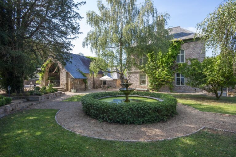 A round fountain in a hedged bed on the lawn, with the house and the arched stone barn behind, Tan House Newland