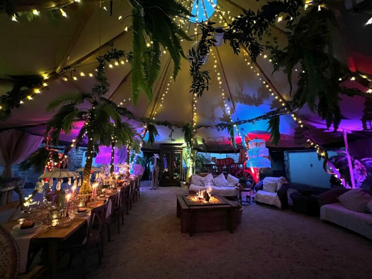 Inside the teepee at night, with fairy lights, palms, coloured uplighting, a long table and the central fire pit, Tan House Newland