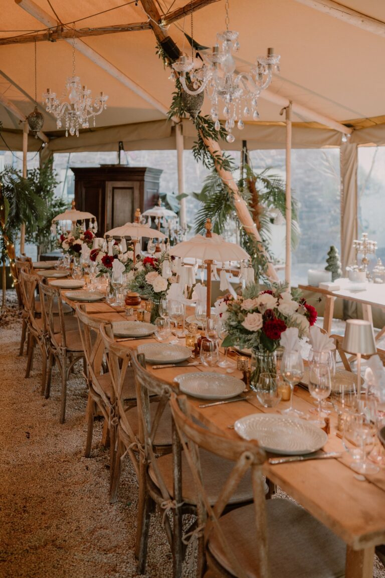 A long trestle table under the teepee laid for a wedding meal with red and white roses, glassware and crystal chandeliers, Tan House Newland
