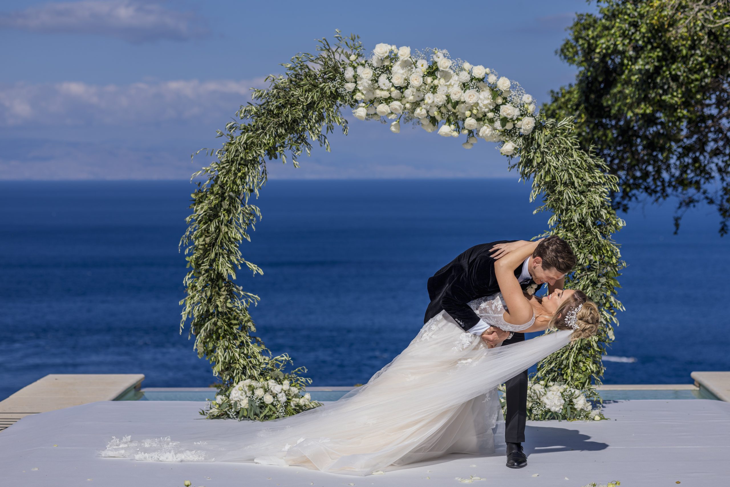 The couple kiss under a circular olive and white rose arch above the Ionian Sea, Taormina Weddings