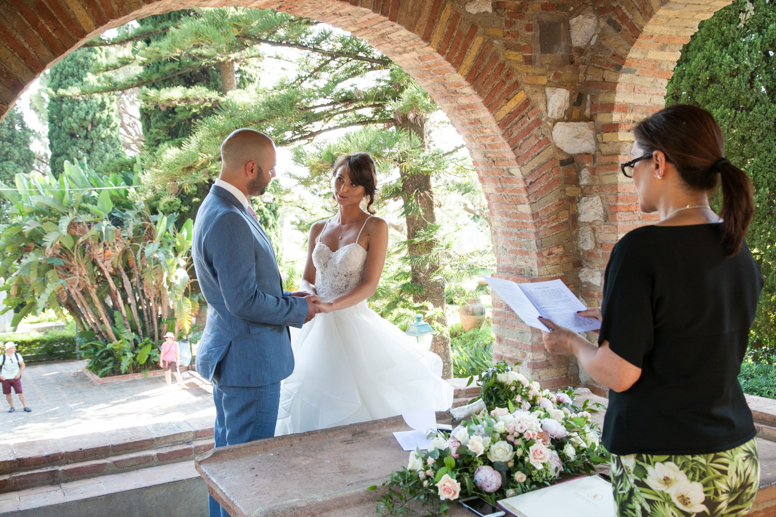 A civil ceremony under a brick arch in a garden in Taormina, the celebrant reading, Taormina Weddings
