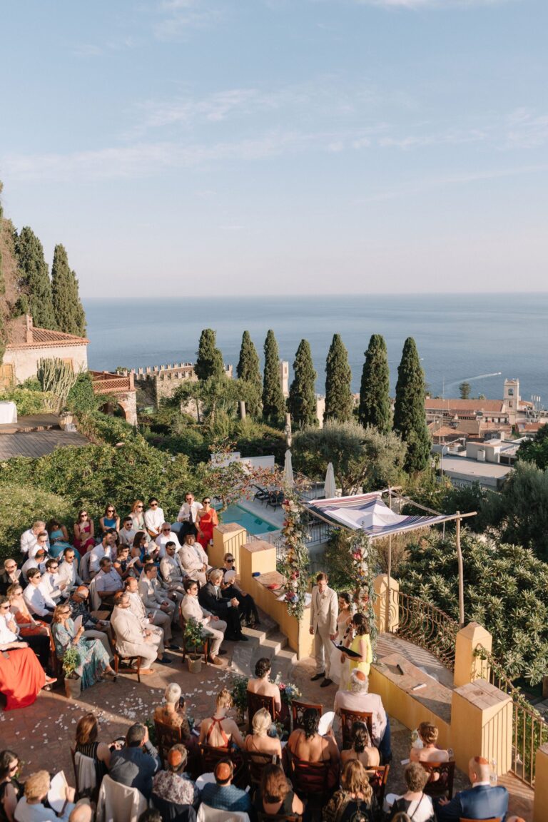 A chuppah on a terrace above the sea, guests seated, the old town and cypresses beyond, Taormina Weddings