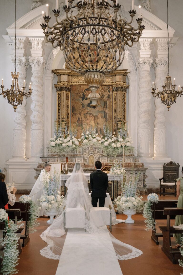 A church ceremony under a large chandelier, white flowers at the altar and a long lace veil, Taormina Weddings